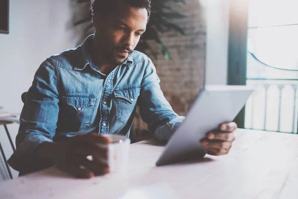 Ernsthafter junger afrikanischer Mann liest Weltnachrichten per digitalem Tablet, während er an einem sonnigen Morgen am Tisch sitzt. Konzept von Coworking People, die zu Hause arbeiten.. — Stockfoto