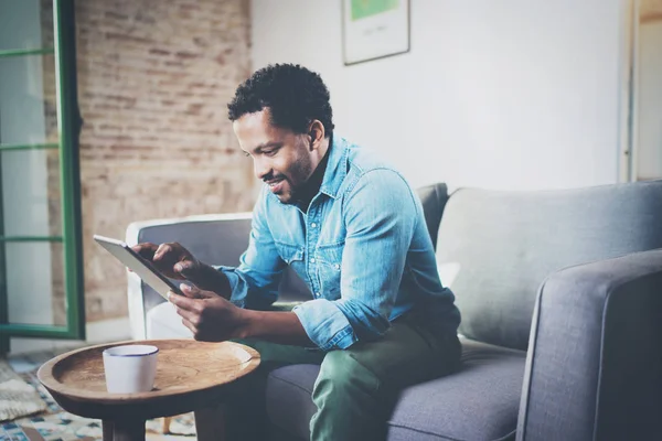 Atractivo empresario africano barbudo usando tableta mientras está sentado en el sofá en su casa moderna.Concepto de los jóvenes que trabajan dispositivos móviles.Fondo borroso . —  Fotos de Stock