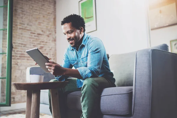 Geconcentreerd bebaarde Afrikaanse man zittend op de Bank thuis werken. Het gebruik van digitale tablet voor nieuwe baan zoeken. Concept van de mobiele apparaten van het werk van jonge mensen. Onscherpe achtergrond. — Stockfoto