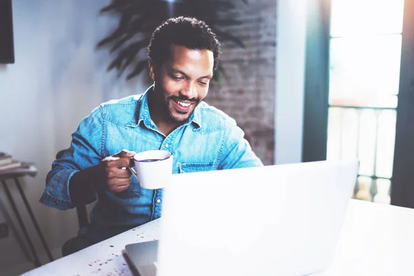 Sonriente hombre africano barbudo que usa el ordenador portátil en casa mientras bebe café negro en la mesa de madera.Concepto de jóvenes que trabajan en dispositivos móviles.Fondo borroso de la ventana . —  Fotos de Stock