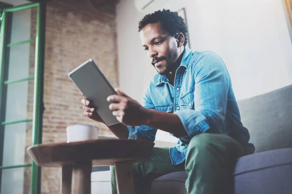 タブレットを使用してビデオ会話近代的なオフィスのソファでリラックスしながら物思いにふけるアフリカ男。若いビジネス人が自宅で仕事のコンセプトです。背景をぼかし。選択と集中. — ストック写真