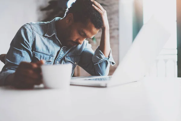 Trött ung afrikansk man använder laptop medan du sitter vid bordet på en solig morgon. Koncept för människor som arbetar hårt hem. Suddig bakgrund, flare effekt. — Stockfoto