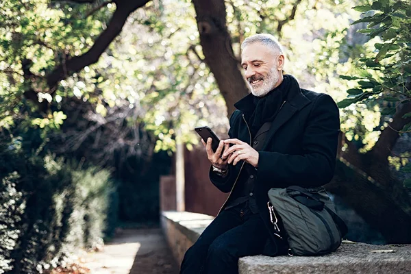 Sorrindo empresário de meia idade usando smartphone enquanto descansa em um parque da cidade.Horizontal, fundo borrado, efeito de filme . — Fotografia de Stock