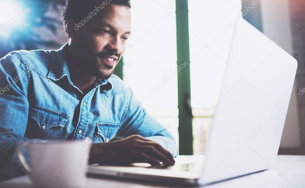 Happy bearded African man using laptop at home while sitting the wooden table.Guy is typing on the notebook keyboard.Concept of young people work mobile devices.Blurred window background,crop.