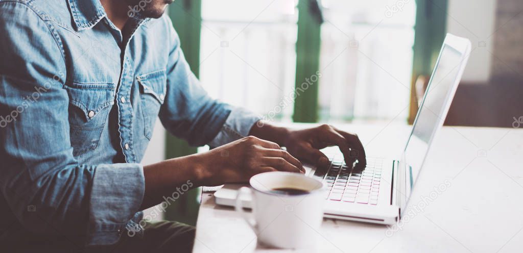 Smiling bearded African man using laptop at home while sitting the wooden table.Male hands typing on the notebook keyboard.Concept of young people work mobile devices.Blurred window background,wide