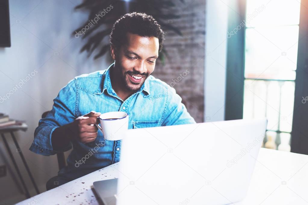 Smiling bearded African man using laptop home while drinking cup black coffee at the wooden table.Concept of young people working on mobile devices.Blurred window background.