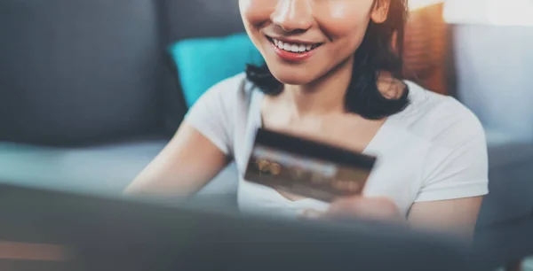 Mujer asiática atractiva feliz usando el ordenador portátil y la tarjeta de crédito de oro para las compras en línea mientras está sentado en el sofá en la sala de estar.Fondo borroso, efecto de bengalas. Amplio . —  Fotos de Stock