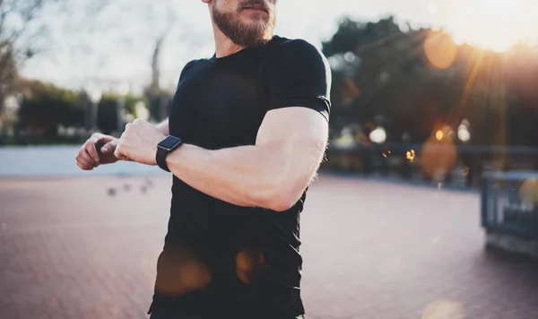 Concepção de estilo de vida de treino ao ar livre.Jovem homem alongando seus músculos do corpo antes do treinamento.Atleta muscular vestindo relógio inteligente na mão exercitando-se fora no parque ensolarado. Fundo desfocado . — Fotografia de Stock