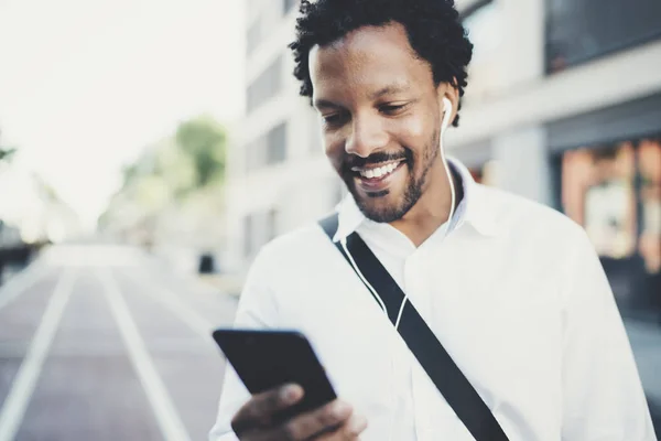 Retrato de un joven africano sonriente usando las manos de un teléfono inteligente mientras está parado en la soleada calle de la ciudad.Concepto de gente de negocios feliz trabajando al aire libre.Fondo borroso . — Foto de Stock
