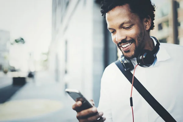 Joven sonriente hombre africano usando el teléfono inteligente en las manos mientras está de pie en la soleada calle de la ciudad.Concepto de gente de negocios feliz trabajando al aire libre.Fondo borroso . —  Fotos de Stock