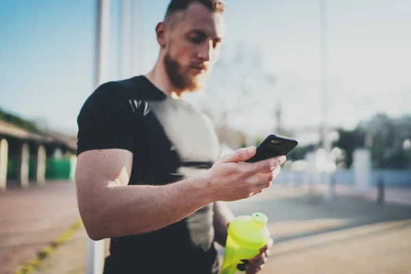 Gespierde knappe atleet controle sport resultaten over smartphone toepassing en slimme horloge na goede training sessie op stadspark in de zonnige ochtend. Onscherpe achtergrond. — Stockfoto