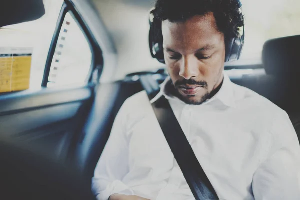 Hombre africano pensativo escuchando música en el teléfono inteligente mientras está sentado en el asiento trasero en el coche de taxista.Concepto de jóvenes felices viajando.Fondo borroso . — Foto de Stock
