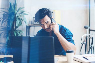 Güneşli ofiste tahta masada otururken çağdaş dizüstü çalışan İspanyol zarif bir iş adamı. Cep telefonundan konuşan adam. Arka plan bulanık. Yatay