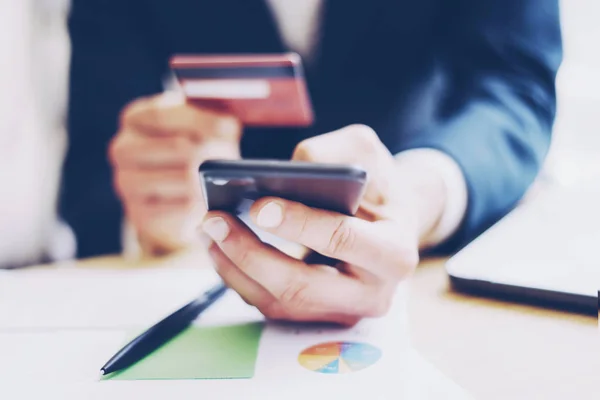 Close-up beeld van zakenman creditcard in de hand te houden en het gebruik van smartphone, papierdocumenten op de houten tafel. Onscherpe achtergrond. Horizontale. — Stockfoto