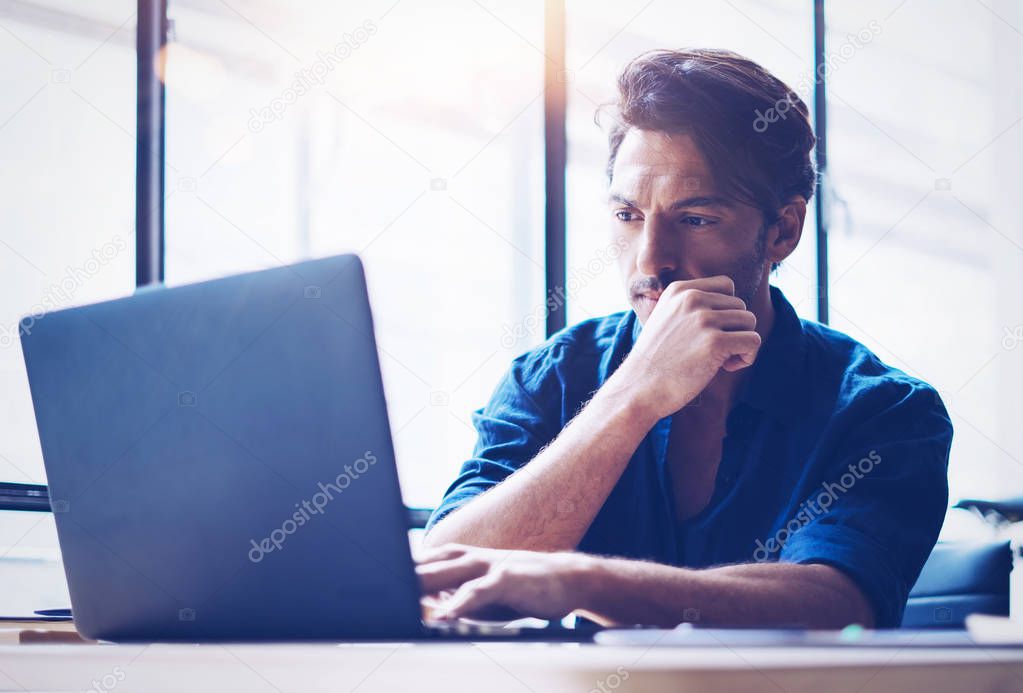 Young handsome banking finance analyst working at sunny office on laptop while sitting at wooden table.Businessman analyze stock reports.Blurred background,horizontal.
