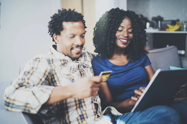 Feliz pareja afroamericana joven sentado en el sofá en casa y de compras en línea a través de la computadora móvil con tarjeta de crédito. Fondo horizontal, borroso . —  Fotos de Stock