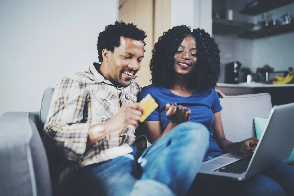 Lyckliga unga afroamerikanska paret sitter på soffan hemma och shoppa online via mobila dator med kreditkort. Horisontell, suddig bakgrund. — Stockfoto