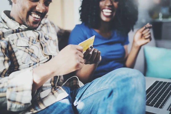 Pasangan muda afrika yang bahagia duduk di sofa di rumah dan berbelanja online melalui komputer seluler dengan kartu kredit. Latar belakang horisontal yang kabur . — Stok Foto