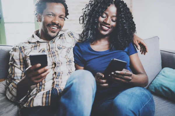 Ευτυχισμένο ζευγάρι Αφρικανικός Αμερικανός χαλαρωτικό μαζί στον καναπέ. Νέοι μαύρο ο άνθρωπος και η φίλη του, χρησιμοποιώντας τα κινητά τηλέφωνα ενώ υπόλοιπα στο σπίτι στο σαλόνι. Οριζόντια, θολή φόντο. — Φωτογραφία Αρχείου