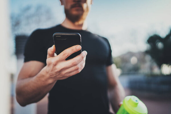 Muscular handsome athlete checking burned calories on smartphone application and smart watch after good workout session on city park in the sunny morning.Blurred background.