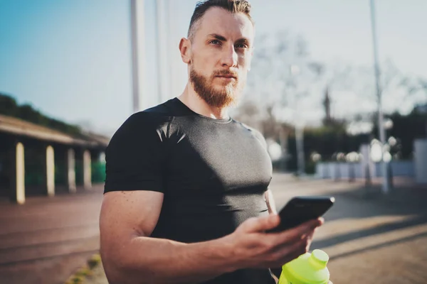 Workout Outdoor-Lifestylekonzept. Junger Mann dehnt seine Armmuskeln vor dem Training. Bärtiger Muskelsportler mit Smartphone und trainiert draußen im sonnigen Park. verschwommener Hintergrund. — Stockfoto