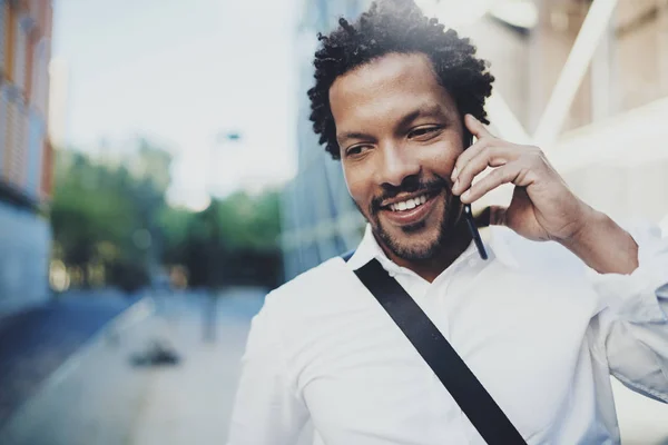 Nahaufnahme Porträt eines lächelnden amerikanisch-afrikanischen Mannes, der sein Smartphone benutzt, um Freunde auf der sonnigen Straße anzurufen.Konzept glücklicher junger gutaussehender Menschen, die Geräte im Freien genießen.verschwommener Hintergrund. — Stockfoto