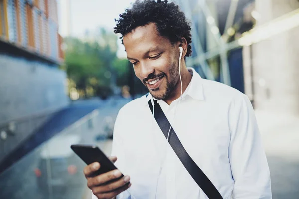 Sonriendo joven hombre afroamericano en auriculares caminando en la ciudad soleada y disfrutando de escuchar música en su teléfono móvil.Fondo borroso.Horizontal . — Foto de Stock