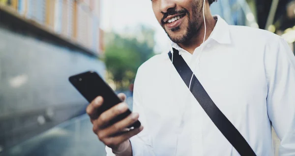 Feliz joven afroamericano hombre en auriculares caminando en la ciudad soleada y disfrutando de escuchar música en su teléfono móvil.Fondo borroso.Horizontal, recortado . —  Fotos de Stock