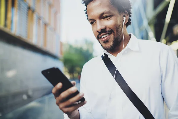 Joven hombre afroamericano en auriculares caminando en la ciudad soleada y disfrutando de escuchar música en su teléfono inteligente.Fondo borroso.Efectos de bengalas. . — Foto de Stock