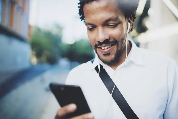 Vista de cerca del joven afroamericano en auriculares caminando por la soleada ciudad y disfrutando de escuchar música en su teléfono inteligente.Fondo borroso.Efectos de bengalas. . —  Fotos de Stock