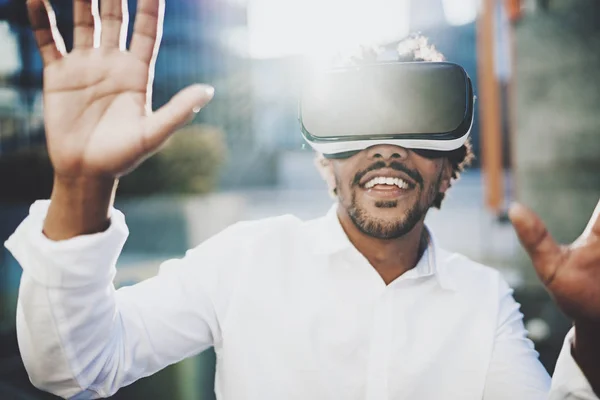 Hombre africano americano barbudo feliz disfrutando de gafas de realidad virtual auriculares o espectáculos 3d mientras camina fuera. Fondo borroso. Horizontal . — Foto de Stock