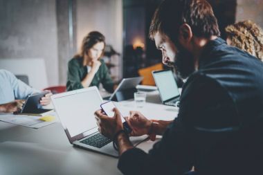 Bearded young coworker working at night office.Man using contemporary laptop and typing on modern smartphone.Horizontal.Blurred background. clipart