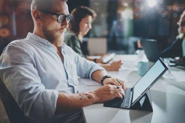 Bärtige junge Kollegen arbeiten in der Nacht office.man mit zeitgenössischen mobilen digitalen tablet.horizontal.unscharfen Hintergrund. Fackeln und Reflexionen. — Stockfoto