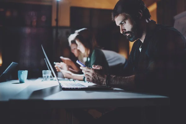 Bärtige junge Mitarbeiter arbeiten in der Nacht Büro. Mann mit zeitgenössischem Laptop und modernen smartphone.horizontal.unscharfen Hintergrund. Fackeln und Reflexionen. — Stockfoto
