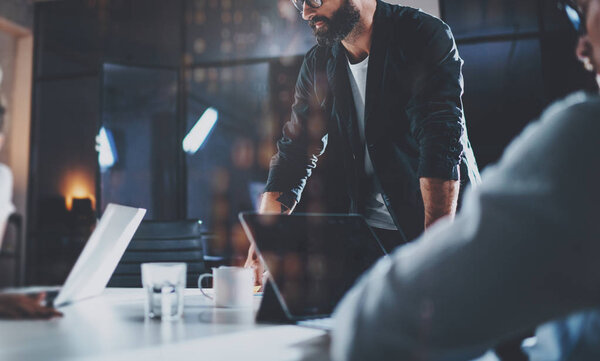 Bearded young coworker working at night office with partners.People using contemporary laptop and modern smartphone.Horizontal.Blurred background.Cropped.