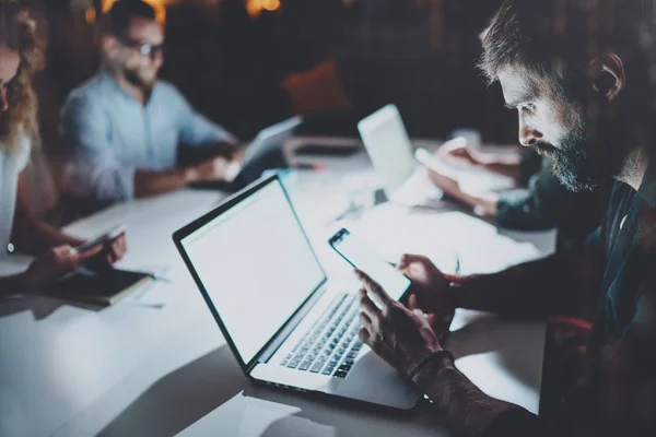 Skäggig ung man arbetar på natten kontor med partners. Personer som använder modern laptop och moderna smartphones. Horizontal.Blurred bakgrund. — Stockfoto