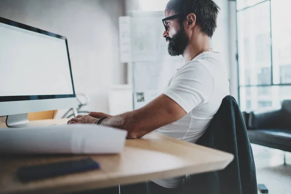 Young bearded professional architect wearing eye glasses working at modern loft studio-office with desktop computer.White blank display screen.Blurred background. Horizontal. — Stock Photo, Image