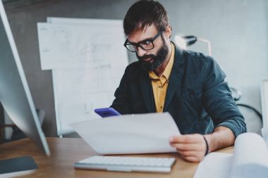 Ahşap masada otururken bilgisayarda lightful ofisinde çalışan genç iş arkadaşınız. Adam ellerini ve onun smartphone kontrol belgesi analiz. Arka plan bulanık. Yatay.