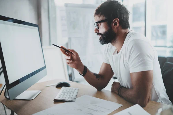 Ung intelligent skäggig man bär glasögon som arbetar på moderna loft studio-kontor med stationär dator. Suddig bakgrund. Horisontell. — Stockfoto