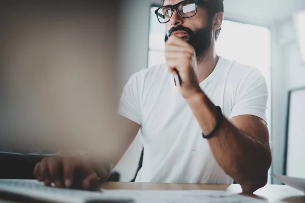 Pensive bearded coworker wearing eye glasses and working at modern loft studio-office.Blurred background. Horizontal. — Stock Photo, Image