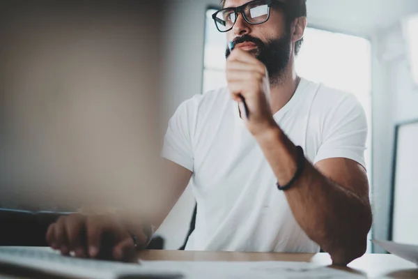 Pensive bearded colleorker wearing eye glasses and working at modern loft studio-office. Blurred background. Горизонталь . — стоковое фото