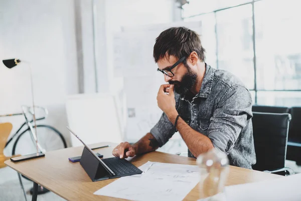 Concepteur barbu pensif travaillant au loft de bureau moderne.Travailleur utilisant ordinateur tablette tactile électronique sur un lieu de travail ensoleillé. Fond flou . — Photo