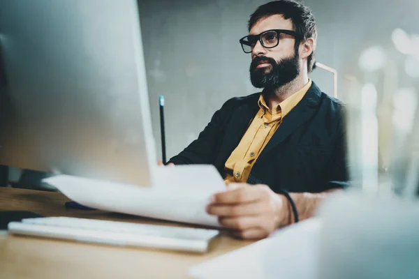 Ung skäggig man som arbetar på soliga kontor på stationär dator medan du sitter på träbord. Affärsman som bär glasögon och casual kläder. Suddig bakgrund, horisontell. — Stockfoto