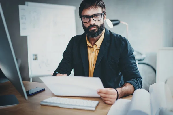 Schöner Architekt, der im sonnigen Büro arbeitet. Bärtiger junger Mann mit Dokument und Blick in die Kamera.Horizontaler, verschwommener Hintergrund. — Stockfoto