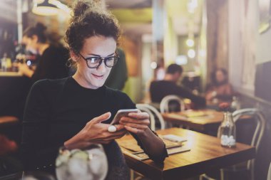 Sevimli kadın güzel gülümsemeyle kafede dinlenme sırasında cep telefonu kısa mesaj okuma. Arka plan bulanık bokeh ve mercek parlamaları etkisi.