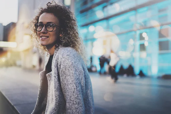 Smiling woman in stylish clothing wearing eye glasses outside in the european night city. Bokeh and flares effect on blurred background. — Stock Photo, Image