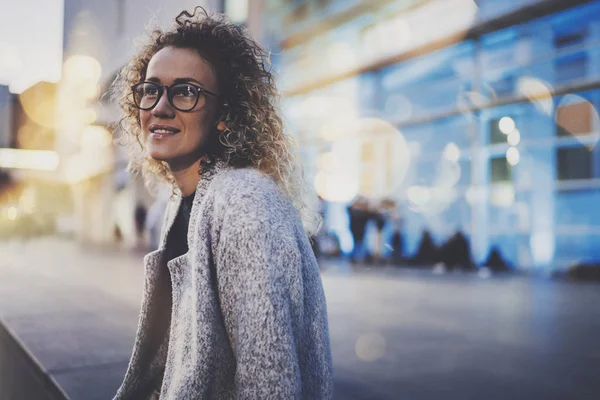 Smiling woman in stylish clothing wearing eye glasses outside in the european night city. Bokeh and flares effect on blurred background. — Stock Photo, Image