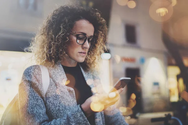 Vrouwelijke hipster dragen rugzak en bril zoeken informatie over mobiele netwerk door slimme telefoon, tijdens lopen in Europese stad op de nacht. Effecten van bokeh en vuurpijlen op onscherpe achtergrond. — Stockfoto