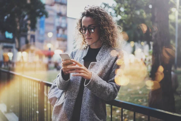 Vrouwelijke hipster dragen rugzak en bril zoeken informatie in mobiele netwerk door slimme telefoon, tijdens de wandeling in Europese stadspark. Effecten van bokeh en vuurpijlen op onscherpe achtergrond. — Stockfoto