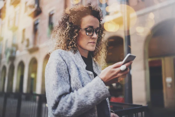 Hansome chica hipster con gafas de búsqueda de información en la red móvil por teléfono inteligente, durante el paseo en la ciudad europea. Efectos de bokeh y bengalas sobre fondo borroso . — Foto de Stock